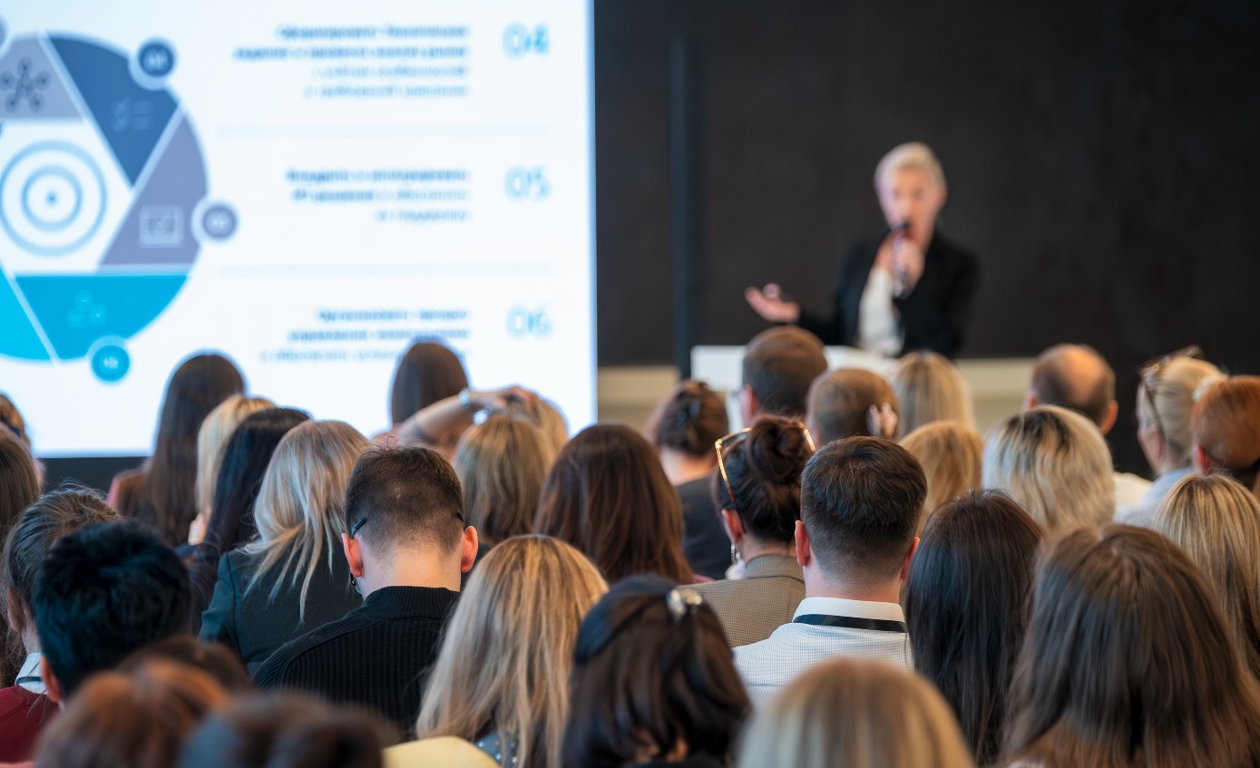 Corporate presenter addressing a large audience at a professional work gathering.